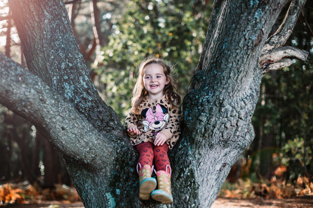 little girl laughing at the camera whilst sat down on a tree branch int he new forest