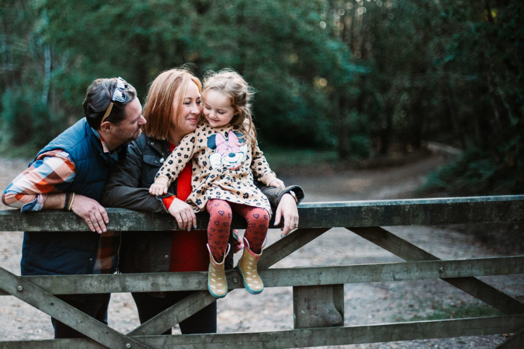 mother and father leaning over a gate inside some woodlands kissing their daughter on the nose
