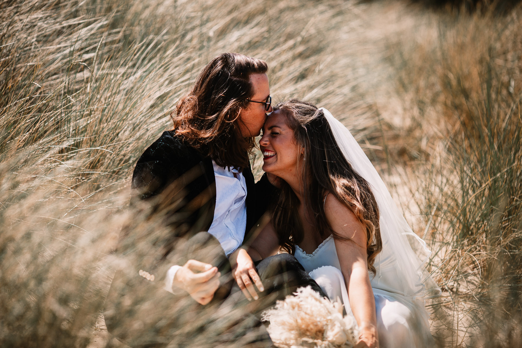 groom sat down with bride kissing her on the forehead in the sand dunes of shell bay beach