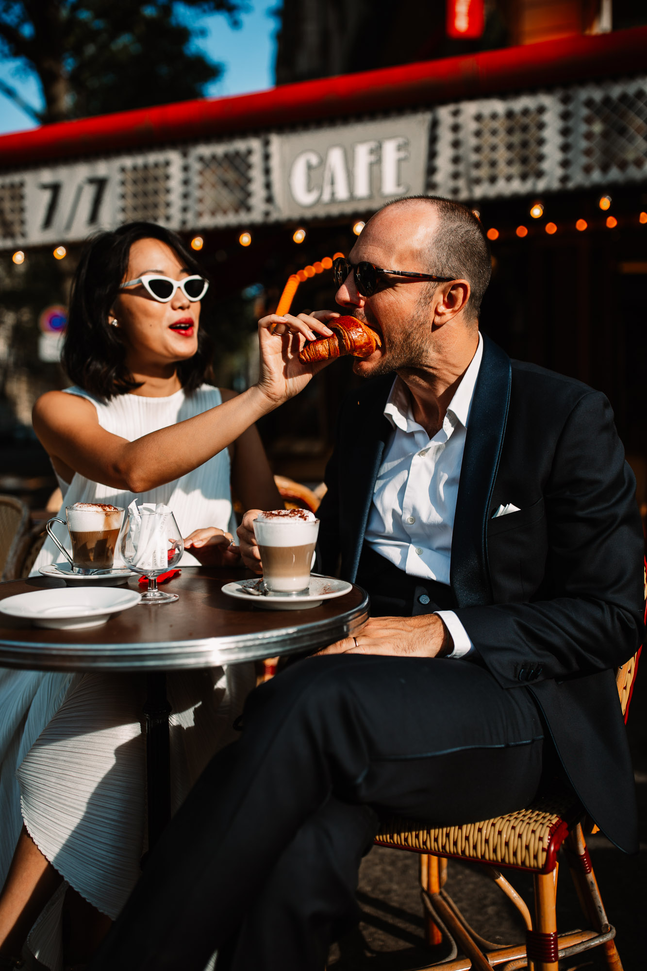 bride wearing sunglasses sat at a terrace of a cafe feeding a croissant to her husband