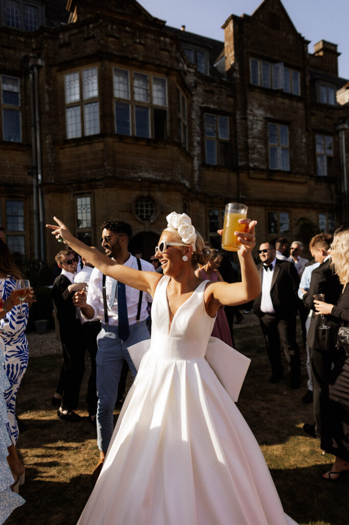 bride with sunglasses on and cocktail in hand dancing in minterne house gardens